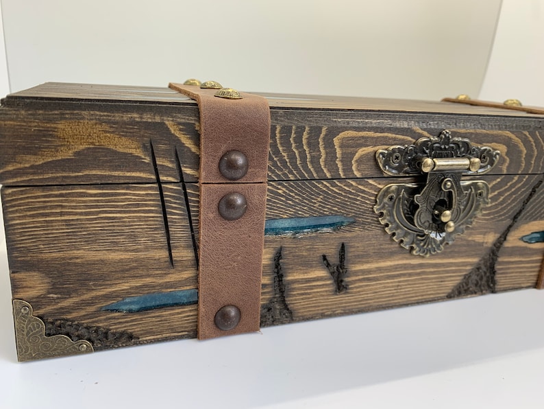May include: A wooden box with a brown leather strap and metal accents. The box has a carved design and a blue resin inlay.