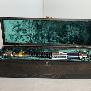 Pode incluir: Um sabre de luz preto com uma pega dourada e detalhes prateados &eacute; apresentado numa caixa de madeira forrada com veludo verde.