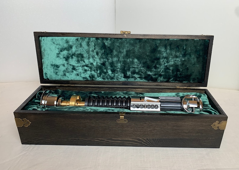 Pode incluir: Um sabre de luz preto com uma pega dourada e detalhes prateados &eacute; apresentado numa caixa de madeira forrada com veludo verde.