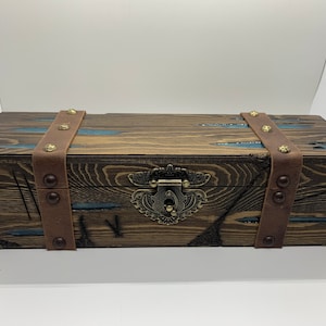 May include: A wooden box with a brown leather strap and metal accents. The box has a blue and brown stained finish and a metal latch.