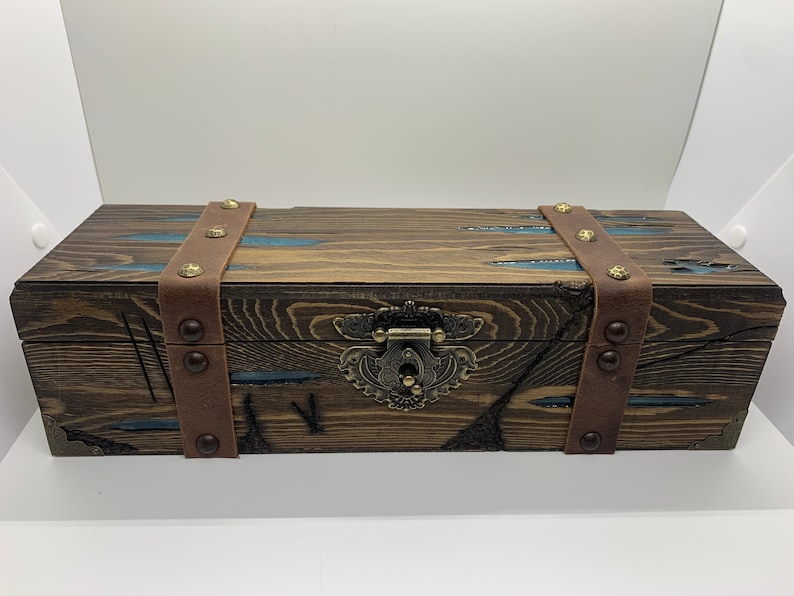 May include: A wooden box with a brown leather strap and metal accents. The box has a blue and brown stained finish and a metal latch.