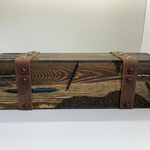 May include: A wooden box with a dark brown stain and leather straps with brass accents. The box has a hinged lid and is closed.