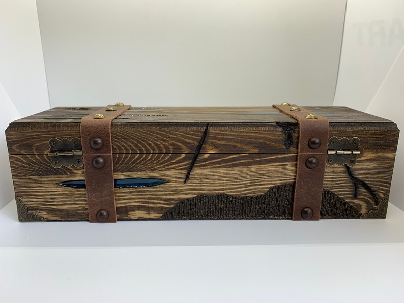 May include: A wooden box with a dark brown stain and leather straps with brass accents. The box has a hinged lid and is closed.