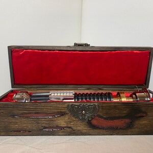 May include: A wooden box with a red velvet lining, containing a silver and black lightsaber. The box has a decorative metal clasp and a burnt wood finish.