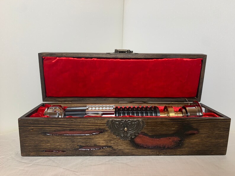 May include: A wooden box with a red velvet lining, containing a silver and black lightsaber. The box has a decorative metal clasp and a burnt wood finish.