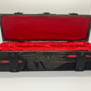 K&ouml;nnte beinhalten: Eine Holzkiste mit rotem Samt-Innenfutter und einem Metallverschluss. Die Kiste hat eine dunkelbraune Holzoptik und ist mit Metallverzierungen verziert.