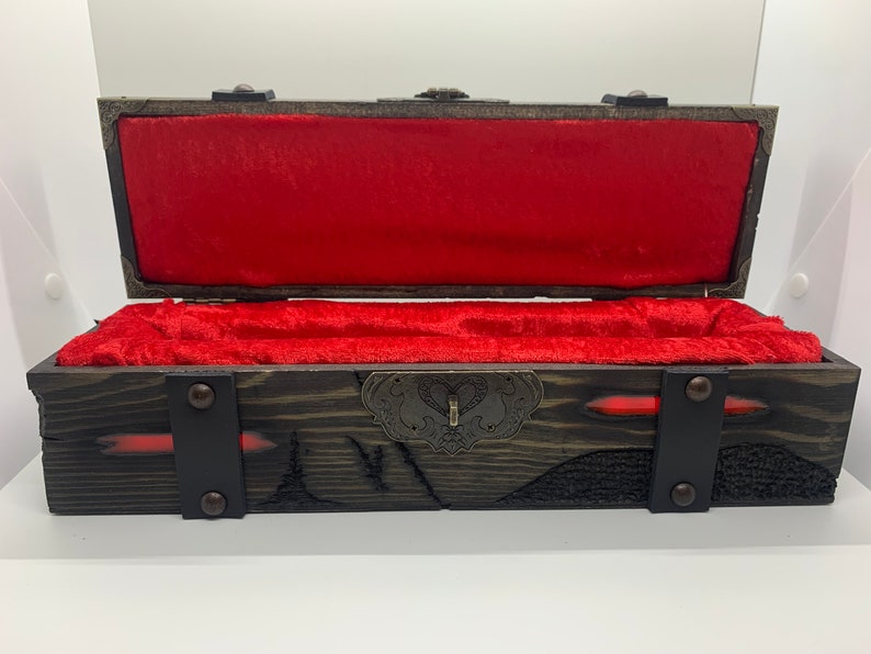 K&ouml;nnte beinhalten: Eine Holzkiste mit rotem Samt-Innenfutter und einem Metallverschluss. Die Kiste hat eine dunkelbraune Holzoptik und ist mit Metallverzierungen verziert.