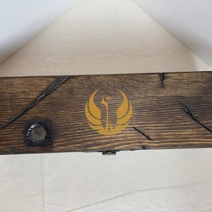 K&ouml;nnte beinhalten: Eine Holzkiste mit einer dunkelbraunen Beize und einem goldenen Rebellenallianz-Symbol auf dem Deckel.