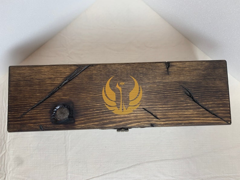 K&ouml;nnte beinhalten: Eine Holzkiste mit einer dunkelbraunen Beize und einem goldenen Rebellenallianz-Symbol auf dem Deckel.
