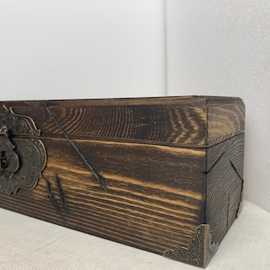 K&ouml;nnte beinhalten: Eine dunkelbraune Holzkiste mit einem Messingverschluss und dekorativen Eckverzierungen. Die Kiste hat eine abgenutzte Oberfl&auml;che und ist aus Holzbrettern gefertigt.