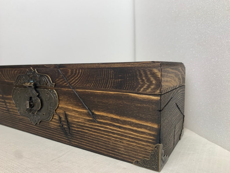 K&ouml;nnte beinhalten: Eine dunkelbraune Holzkiste mit einem Messingverschluss und dekorativen Eckverzierungen. Die Kiste hat eine abgenutzte Oberfl&auml;che und ist aus Holzbrettern gefertigt.