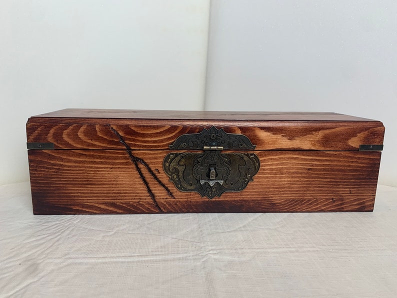 May include: A wooden box with a dark brown stain and a decorative metal latch. The box has a rustic look and is made of natural wood.