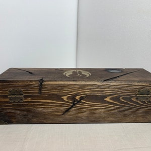 K&ouml;nnte beinhalten: Eine Holzkiste mit einer dunkelbraunen Beize. Die Kiste hat zwei Messing Scharniere und ein dekoratives Design auf der Oberseite.
