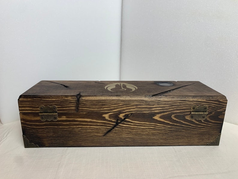 K&ouml;nnte beinhalten: Eine Holzkiste mit einer dunkelbraunen Beize. Die Kiste hat zwei Messing Scharniere und ein dekoratives Design auf der Oberseite.