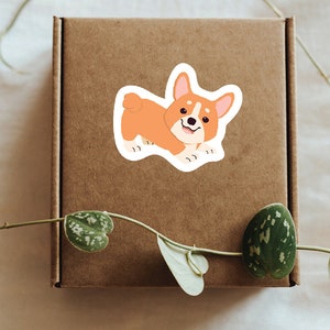 Puede incluir: Una caja de cart&oacute;n marr&oacute;n con una pegatina de un perro corgi de dibujos animados. El corgi es naranja y blanco con una expresi&oacute;n feliz.