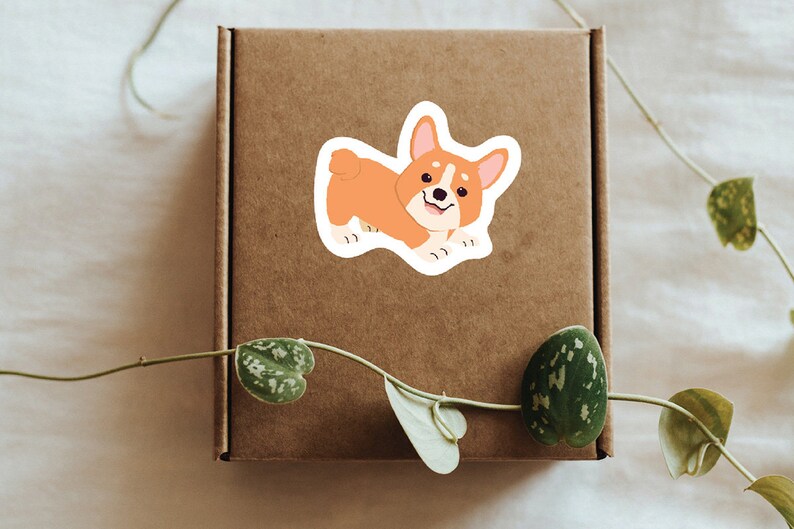Puede incluir: Una caja de cart&oacute;n marr&oacute;n con una pegatina de un perro corgi de dibujos animados. El corgi es naranja y blanco con una expresi&oacute;n feliz.