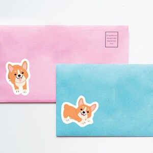 Puede incluir: Dos sobres, uno rosa y otro azul, con una imagen de dibujos animados de un perro corgi en cada uno. El sobre rosa tiene el texto "Entgeltmarken im Inland der Deutschen Post." impreso en &eacute;l.