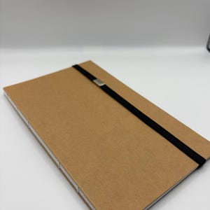 K&ouml;nnte beinhalten: Ein rechteckiges Notizbuch in Kraftpapierfarbe mit einem schwarzen Gummibandverschluss und einem silberfarbenen Metallakzent. Das Notizbuch hat wei&szlig;e N&auml;hte entlang des R&uuml;ckens und der Kanten, vor wei&szlig;em Hintergrund.