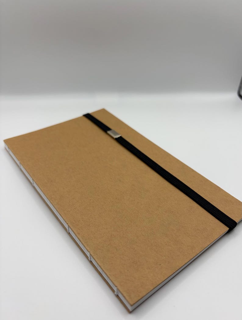 K&ouml;nnte beinhalten: Ein rechteckiges Notizbuch in Kraftpapierfarbe mit einem schwarzen Gummibandverschluss und einem silberfarbenen Metallakzent. Das Notizbuch hat wei&szlig;e N&auml;hte entlang des R&uuml;ckens und der Kanten, vor wei&szlig;em Hintergrund.