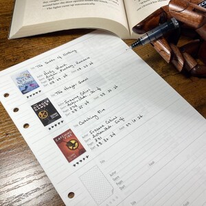 May include: A handwritten book list with titles like "The Queen of Nothing", "The Hunger Games", and "Catching Fire". The list includes author, genre, page count, and start/finish dates. A wooden hand holds a pen, and an open book is in the background.