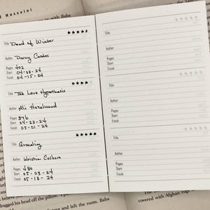 May include: A close-up of a white notebook with a black pen. The notebook is divided into sections for recording book titles, authors, pages, start date, and finish date. The notebook is filled with handwritten entries for books read, including "The Dead of Winter" by Darcy Coates, "The Love Hypothesis" by Ali Hazelwood, and "Graceling" by Kristin Cashore.