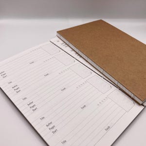 K&ouml;nnte beinhalten: Zwei handgefertigte Journale mit einem braunen Kraftpapier-Einband und einer gen&auml;hten Bindung. Die offenen Journalseiten haben ein liniertes Layout mit Feldern f&uuml;r Titel, Autor, Seiten und Start-/Enddaten. Die Journale sind zum Schreiben.