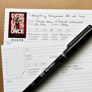 Peut inclure: Une fiche de critique de film pour "Everything Everywhere All at Once" avec l'affiche du film, le titre, le réalisateur, la durée et l'année de sortie. Un stylo noir est posé sur la carte, avec le mot "PILOT" visible.