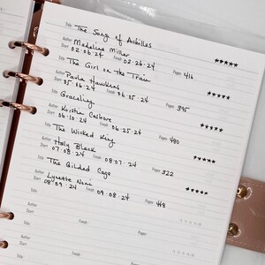 May include: A close-up of a planner with a list of books to read, including titles like "The Song of Achilles", "The Girl on the Train", "Gracefully", "The Wicked King", and "The Gilded Cage". The planner has a pink leather cover and gold rings.