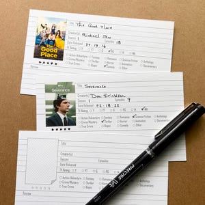 May include: Three white index cards with handwritten details and images from the TV shows "The Good Place" and "Severance." A black pen rests on the cards. The cards are on a brown surface.