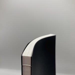 K&ouml;nnte beinhalten: Ein handgen&auml;htes schwarzes Notizbuch mit gew&ouml;lbtem R&uuml;cken. Die Seiten sind wei&szlig;, und die Bindung ist mit beigem Faden sichtbar. Der Einband ist einfarbig schwarz, und das Notizbuch ist vor einem hellgrauen Hintergrund platziert.