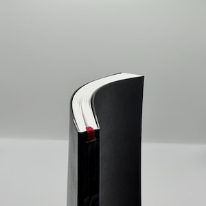 K&ouml;nnte beinhalten: Ein schwarzes Hardcover-Notizbuch mit wei&szlig;en Papierseiten. Ein rotes Band als Lesezeichen ist zu sehen. Das Notizbuch wird von der Seite gezeigt, mit leicht gew&ouml;lbten Seiten.