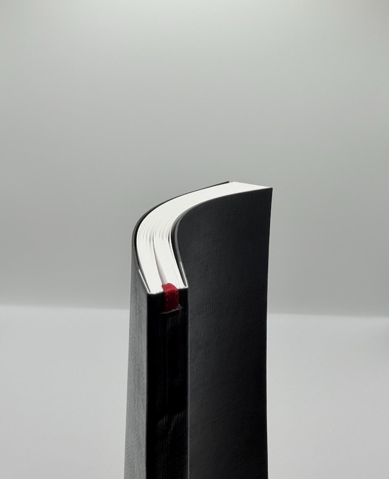 K&ouml;nnte beinhalten: Ein schwarzes Hardcover-Notizbuch mit wei&szlig;en Papierseiten. Ein rotes Band als Lesezeichen ist zu sehen. Das Notizbuch wird von der Seite gezeigt, mit leicht gew&ouml;lbten Seiten.