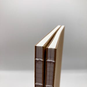 K&ouml;nnte beinhalten: Zwei handgefertigte Notizb&uuml;cher mit Kraftpapier-Einb&auml;nden und wei&szlig;er, gen&auml;hter Bindung. Die Seiten sind strahlend wei&szlig; und die Einb&auml;nde hellbraun. Die Notizb&uuml;cher liegen nebeneinander, die R&uuml;cken zum Betrachter.