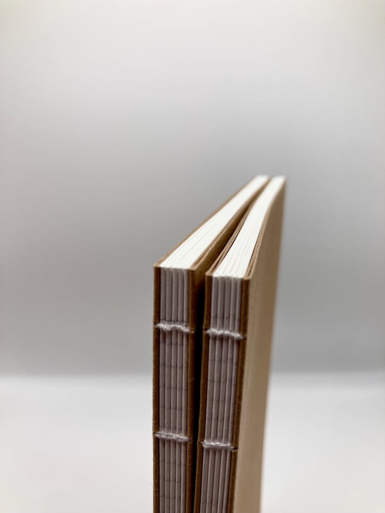 K&ouml;nnte beinhalten: Zwei handgefertigte Notizb&uuml;cher mit Kraftpapier-Einb&auml;nden und wei&szlig;er, gen&auml;hter Bindung. Die Seiten sind strahlend wei&szlig; und die Einb&auml;nde hellbraun. Die Notizb&uuml;cher liegen nebeneinander, die R&uuml;cken zum Betrachter.