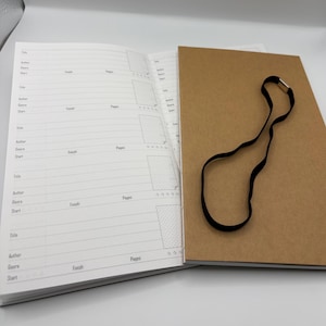 K&ouml;nnte beinhalten: Ein offenes Notizbuch mit linierten Seiten und einem braunen Einband. Die linke Seite hat Abschnitte f&uuml;r Titel, Autor, Genre und Start/Ende. Ein schwarzes Band mit goldenem Verschluss ist befestigt.