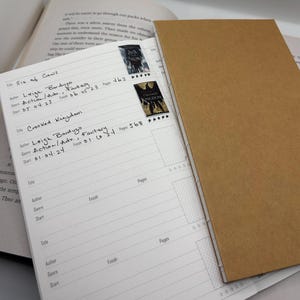 K&ouml;nnte beinhalten: Ein offenes Buchjournal mit braunem Einband zeigt eine linierte Seite mit handschriftlichen Eintr&auml;gen und Buchaufklebern. Die Eintr&auml;ge umfassen Buchtitel, Autor, Genre und Seitenzahlen. Zwei Buchaufkleber sind sichtbar.