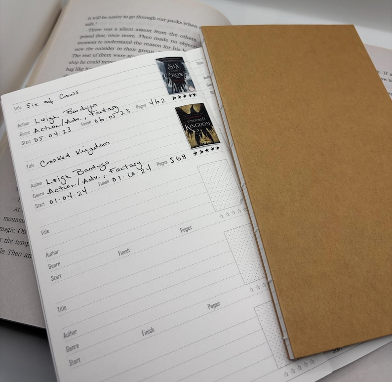 K&ouml;nnte beinhalten: Ein offenes Buchjournal mit braunem Einband zeigt eine linierte Seite mit handschriftlichen Eintr&auml;gen und Buchaufklebern. Die Eintr&auml;ge umfassen Buchtitel, Autor, Genre und Seitenzahlen. Zwei Buchaufkleber sind sichtbar.