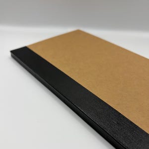 May include: A rectangular notebook featuring a tan, smooth cover and a thick, textured black spine. The notebook is positioned at an angle, highlighting the contrast between the light brown paper surface and the dark, woven-like black binding.