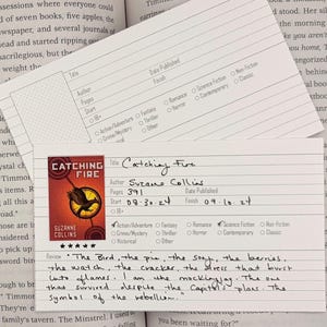 Puede incluir: Una tarjeta de lectura de "Catching Fire" de Suzanne Collins, con una imagen de un pájaro. La tarjeta incluye notas manuscritas y detalles sobre el libro, como las fechas de inicio y fin, y el número de páginas.
