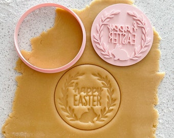 May include: Pink plastic cookie cutters with a wreath design and the text "Happy Easter". The cutters are on top of a sheet of unbaked cookie dough.