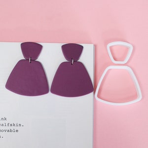 Könnte beinhalten: Ein Paar violette geometrische Ohrringe mit einem silbernen Hakenverschluss. Die Ohrringe liegen auf einer weißen Oberfläche neben einem weißen Plastik-Ausstecher in Form eines Kleides.