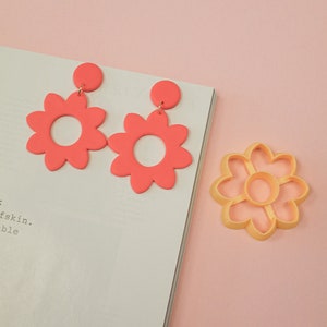 Op de afbeelding: Een paar roze bloemvormige oorbellen met een gouden haak. De oorbellen liggen op een wit oppervlak naast een gele bloemvormige koekjesuitsteker.