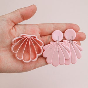 Può includere: Un paio di orecchini rosa a forma di conchiglia con un tagliapasta a forma di conchiglia abbinato. Gli orecchini sono realizzati in pasta polimerica e hanno un aspetto delicato e femminile.