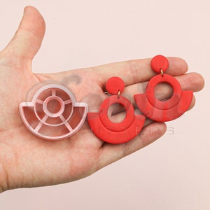 Può includere: Un paio di orecchini in argilla polimerica rossa con un design geometrico. Gli orecchini sono realizzati con due cerchi e una semicirconferenza. Gli orecchini sono mostrati con uno stampo in plastica rosa per la realizzazione degli orecchini.