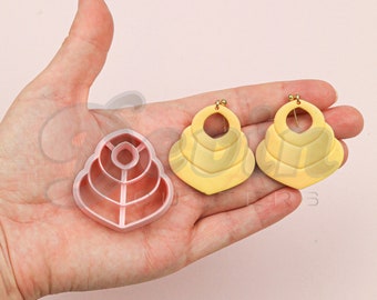 Può includere: Un tagliapasta in plastica rosa a forma di design geometrico a strati e due orecchini gialli con lo stesso design geometrico a strati.