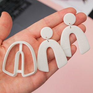Peut inclure: Trois boucles d'oreilles en argile polymère blanche en forme d'arche. Les boucles d'oreilles sont de tailles différentes et ont une finition scintillante.