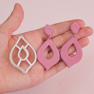 Puede incluir: Un par de pendientes de color rosa en forma de lágrima con una superficie texturizada. Los pendientes se muestran con un molde de plástico blanco para hacer los pendientes.