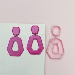 May include: A pair of pink geometric earrings with a cutout design. The earrings are made of polymer clay and are lightweight and comfortable to wear.