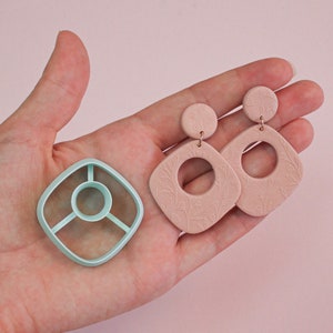 May include: A pair of pink polymer clay earrings with a floral design and a square shape. The earrings are shown on a hand holding a light blue plastic mold.