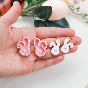 May include: A pair of pink bunny-shaped silicone molds and a pair of white bunny-shaped earrings with pink hearts. The earrings have gold hooks.